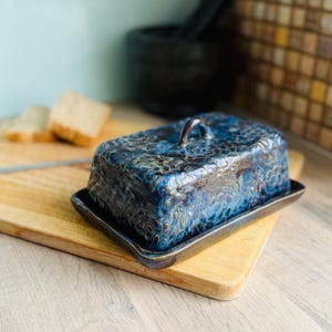 May include: A blue ceramic butter dish with a lid, featuring a textured floral pattern. The dish sits on a rectangular base and has a small handle on top. The butter dish is placed on a wooden chopping board.