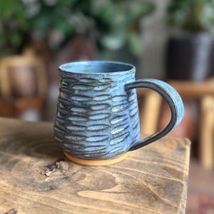 May include: A handmade ceramic mug with a textured, oval pattern in shades of blue and brown. The mug has a rounded handle and sits on a light brown base. The mug is displayed on a wooden surface.