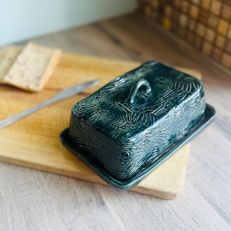 May include: A dark green ceramic butter dish with a rectangular base and a matching lid featuring a textured, leaf-like pattern and a small arched handle on top. The glaze has a glossy finish that highlights the intricate, embossed surface details.
