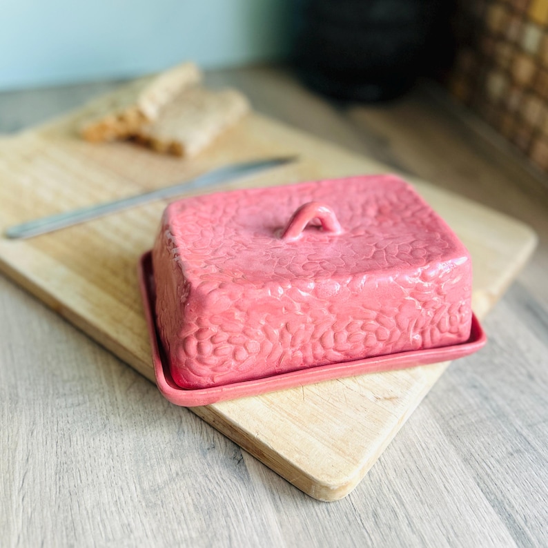 May include: A rectangular pink ceramic butter dish with a textured, pebble-like surface pattern and a small arched handle on the lid. The dish consists of a matching base tray and a removable cover, featuring a consistent matte pink finish throughout.