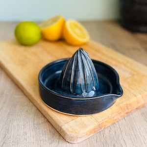 May include: A dark blue ceramic citrus juicer with a ridged cone and a wide bowl. The juicer is on a wooden chopping board, with a lime and halved lemons in the background. A kitchen tool for making fresh juice.