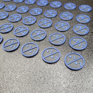 Pode incluir: Uma coleção de pequenas fichas circulares cinzentas com bordas azuis. Cada ficha é dividida em duas secções com números e uma linha no centro. Os números incluem 1, 2, 3 e 4. As fichas estão dispostas numa superfície texturizada.
