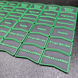 May include: A collection of gray and green game tokens. Each token has a gray base with a green border and text. The text includes words like "First Strike", "Haste", and "Indestructible". The tokens are arranged in a grid pattern.