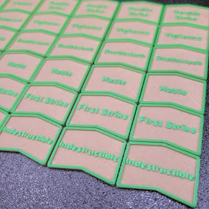 May include: A collection of beige game tokens with green borders and text. The tokens are arranged in rows and columns, with the words "First Strike", "Indestructible", "Haste", and other game terms visible.