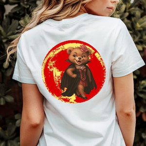 Könnte beinhalten: Weißes T-Shirt mit einem Cartoon-Bären, der als Vampir verkleidet ist. Der Bär trägt einen schwarzen Umhang und ein rotes Kleid mit einer Rose im Haar. Das Design befindet sich auf einem roten und gelben kreisförmigen Hintergrund.