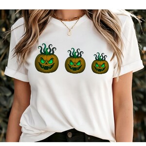 Könnte beinhalten: Weißes T-Shirt mit drei Halloween-Kürbis-Designs. Jeder Kürbis hat ein geschnitztes Gesicht mit leuchtend grünen Augen und einem grinsenden Mund. Die Kürbisse sind in verschiedenen Größen, mit grünen, lockigen Ranken oben.