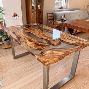 Może przedstawiać: Prostokątny stół jadalny z przezroczystym blatem z żywicy epoksydowej, prezentujący naturalne wzory słojów drewna. Stół wspierają nogi ze szczotkowanego metalu. Drewno ma ciepły brązowy odcień.