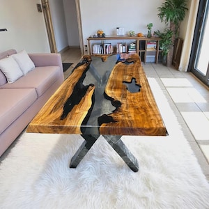 May include: A rectangular wooden coffee table with a clear epoxy river running through the center. The table has a natural wood grain finish with black accents and sits on a black metal X-shaped base. The table measures approximately 72 inches long.