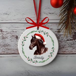 May include: Circular white Christmas ornament with a brown horse wearing a Santa hat. The ornament is decorated with a green wreath and red berries, with the word "Beauty". A red ribbon is attached for hanging.