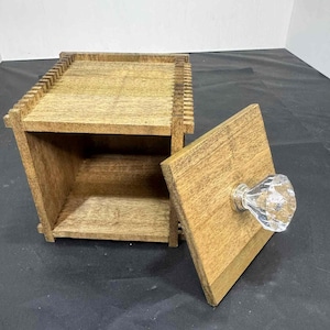 May include: Wooden box with a hinged lid and a clear crystal knob. The box is made of light brown wood and has a rectangular shape. The lid is open, revealing the interior of the box.
