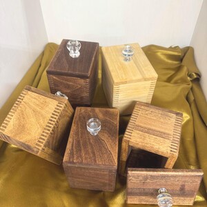 May include: Six wooden boxes with clear glass knobs. The boxes are various shades of brown and natural wood, with a rectangular shape. The boxes are arranged on a gold-colored fabric.