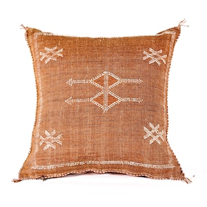 May include: Square, rust-colored decorative pillow with a geometric design. The pillow features a woven texture and is adorned with white embroidered patterns. The central design is a diamond shape with arrow-like extensions. Tassels are visible at the corners.