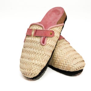 Może przedstawiać: Para plecionych chodaków z rafii z różowymi zamszowymi akcentami. Chodaki mają naturalny beżowy kolor z plecionym wzorem, różowym zamszowym paskiem i czarną podeszwą. Wnętrze jest wyłożone różowym zamszem.