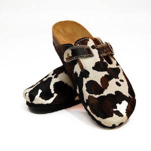 May include: A pair of clogs with a cowhide pattern in black, brown, and white. The clogs have a leather strap with a buckle and a wooden sole. The clogs are designed for comfort and style.
