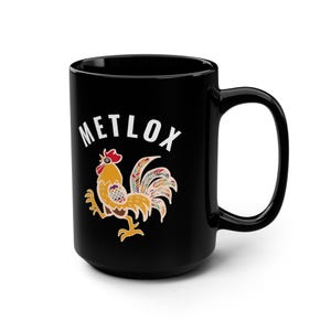May include: Black ceramic mug featuring the word "METLOX" in white. A colourful rooster illustration is below the text. The rooster is yellow, brown, red, and has a multi-coloured tail.