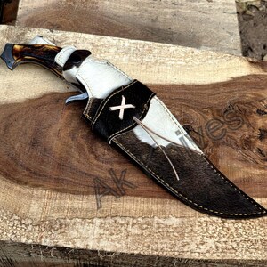 May include: A hunting knife with a brown antler handle and a black and white cowhide sheath. The sheath features a black leather strap with a white cross-stitch design. The knife is resting on a wooden surface.