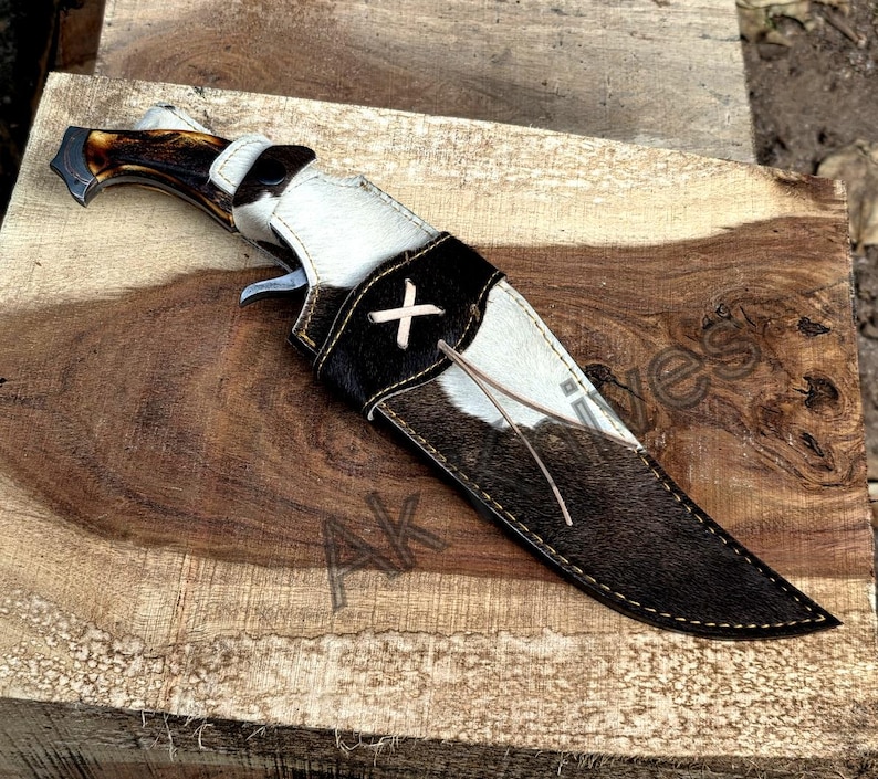 May include: A hunting knife with a brown antler handle and a black and white cowhide sheath. The sheath features a black leather strap with a white cross-stitch design. The knife is resting on a wooden surface.