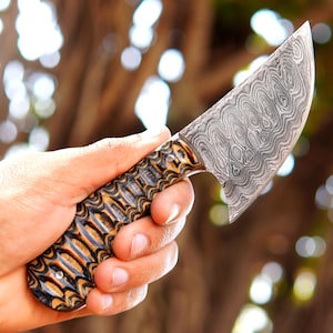 May include: A fixed-blade knife featuring a distinct Damascus steel blade with a wavy, layered pattern. The handle is crafted from textured, multi-colored wood with alternating dark and light brown tones, secured by a single visible metal pin.