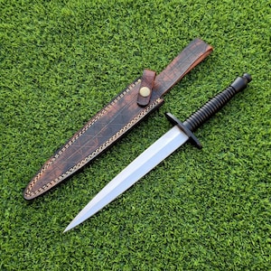 Könnte beinhalten: Ein Dolch mit doppelseitiger Klinge und schwarzem Griff, auf einer grünen Oberfläche. Der Dolch wird von einer braunen Lederscheide mit Knopfverschluss begleitet. Die Scheide hat ein strukturiertes Muster und dekorative Nähte.