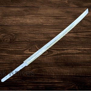 May include: A long, straight, silver-colored sword blade with a pointed tip. The blade has a flat, rectangular profile and is set against a dark brown wooden background. The text "Ak Knives" is visible near the base of the blade.