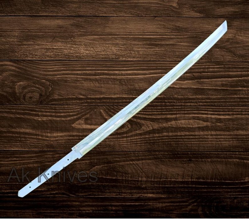 May include: A long, straight, silver-colored sword blade with a pointed tip. The blade has a flat, rectangular profile and is set against a dark brown wooden background. The text "Ak Knives" is visible near the base of the blade.