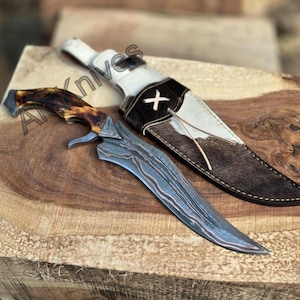 May include: A handcrafted hunting knife with a patterned steel blade and a brown and tan antler handle. The knife rests on a wooden surface next to a leather sheath with a white and brown cowhide design.