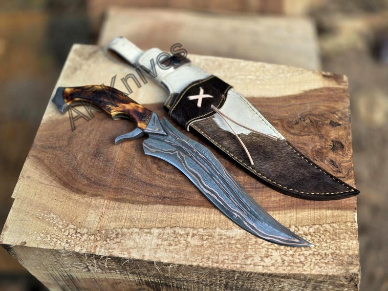 May include: A handcrafted hunting knife with a patterned steel blade and a brown and tan antler handle. The knife rests on a wooden surface next to a leather sheath with a white and brown cowhide design.