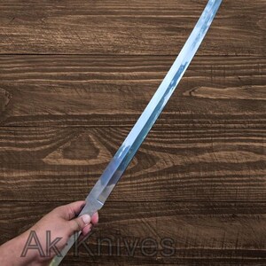 May include: A long, straight, silver-colored blade with a light green handle is held in a hand. The blade reflects the sky. The text "Ak Knives" is visible near the handle. The blade appears to be a knife or sword.