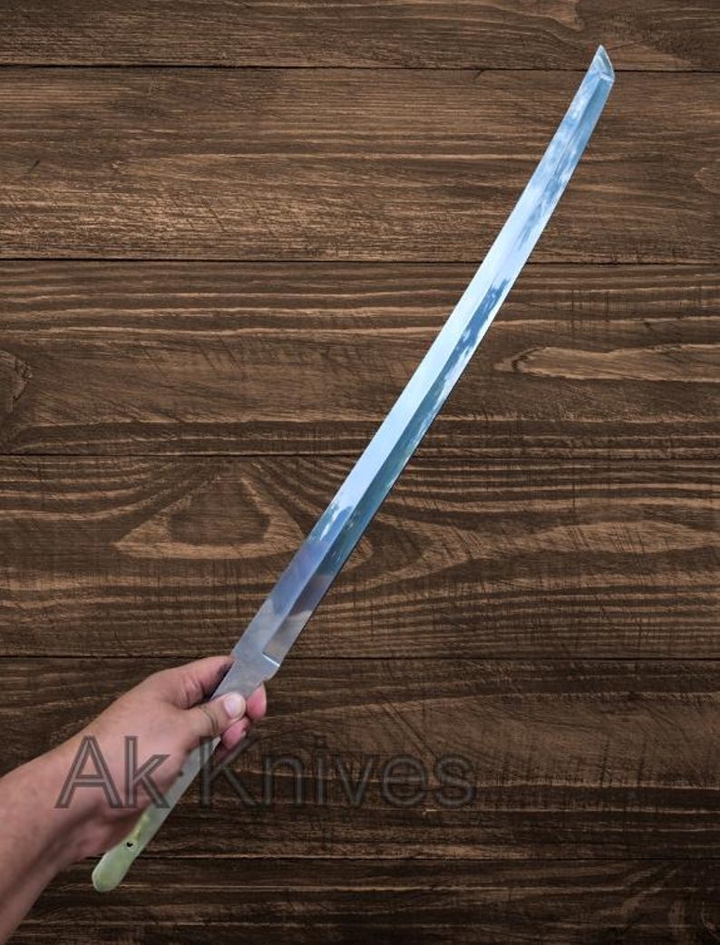 May include: A long, straight, silver-colored blade with a light green handle is held in a hand. The blade reflects the sky. The text "Ak Knives" is visible near the handle. The blade appears to be a knife or sword.