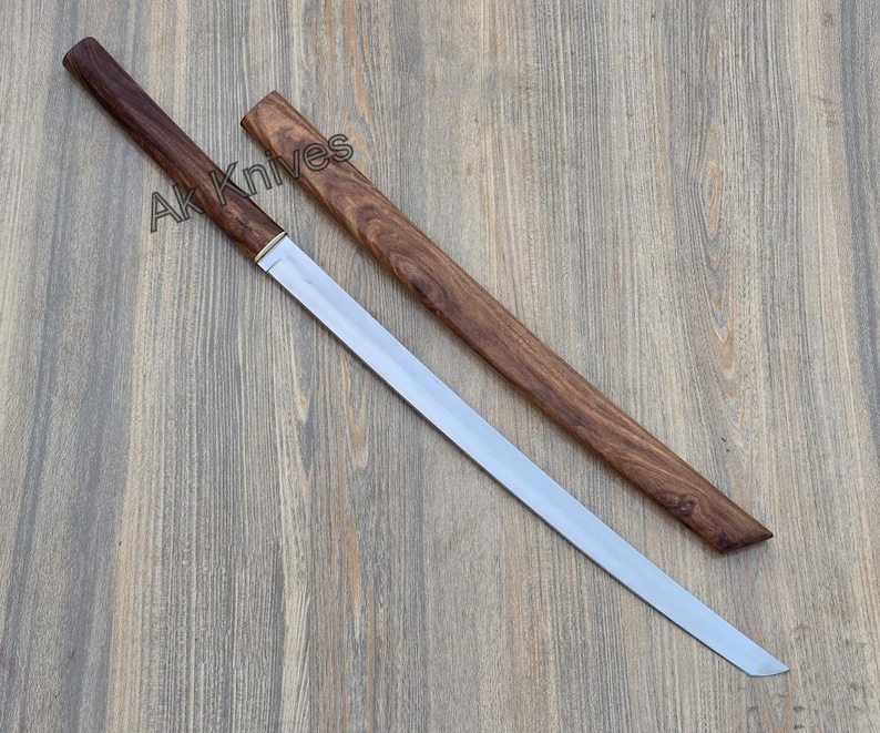 May include: A katana-style sword with a polished steel blade and a dark wood handle and sheath. The text "Ak Knives" is printed on the handle. The sword is displayed on a wooden surface.
