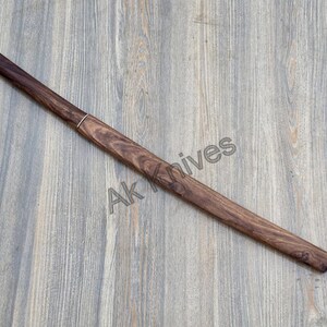 May include: A wooden knife sheath, finished in dark brown, with a silver accent ring. The sheath is long and slender, with an angled tip. The text "Ak Knives" is visible on the surface.