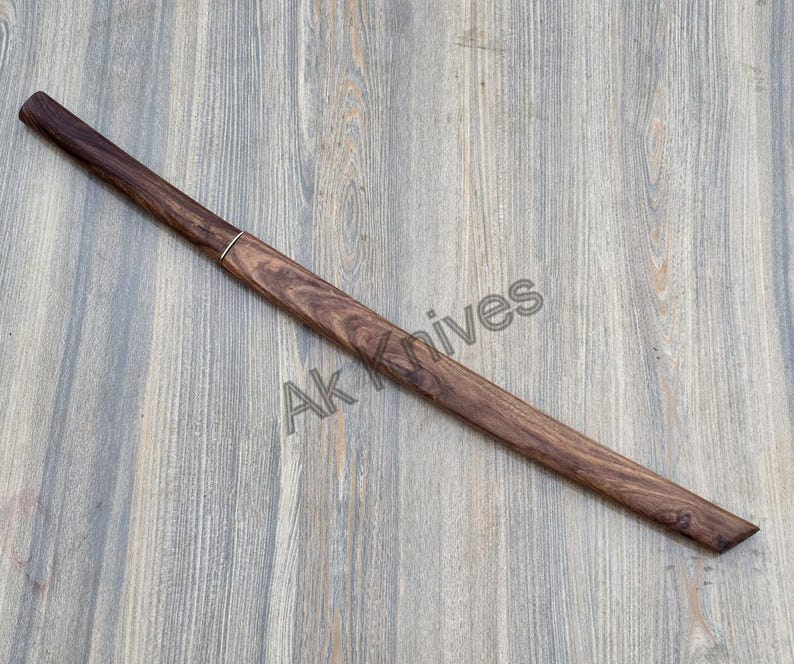 May include: A wooden knife sheath, finished in dark brown, with a silver accent ring. The sheath is long and slender, with an angled tip. The text "Ak Knives" is visible on the surface.