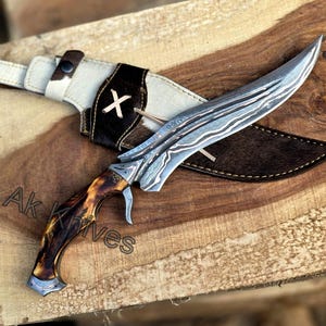 May include: A handcrafted hunting knife with a patterned steel blade and a handle made of brown and tan material. The knife rests on a wooden surface, accompanied by a leather sheath with a white and black cowhide design. The text "Ak Knives" is visible.
