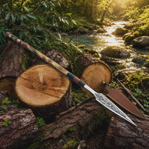 Puede incluir: Una lanza artesanal con un asta de madera y una hoja de metal decorativa. La lanza presenta un agarre envuelto en cuero con detalles en negro, marrón y azul. La hoja tiene un diseño grabado intrincado. La lanza se exhibe sobre un lecho de troncos.