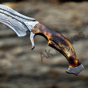 May include: Close-up of a custom-made knife with a curved, patterned blade. The handle is crafted from dark brown material with a unique, textured appearance. The blade has a silver and copper design. The text "Ak Knives" is visible on the handle.