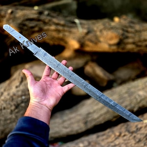 Könnte beinhalten: Ein langes, silberfarbenes Messer mit gemusterter Klinge und Griff. Der Text "Ak Knives" ist auf dem Griff zu sehen. Das Messer wird in einer Hand vor einem verschwommenen, natürlichen Hintergrund gehalten.