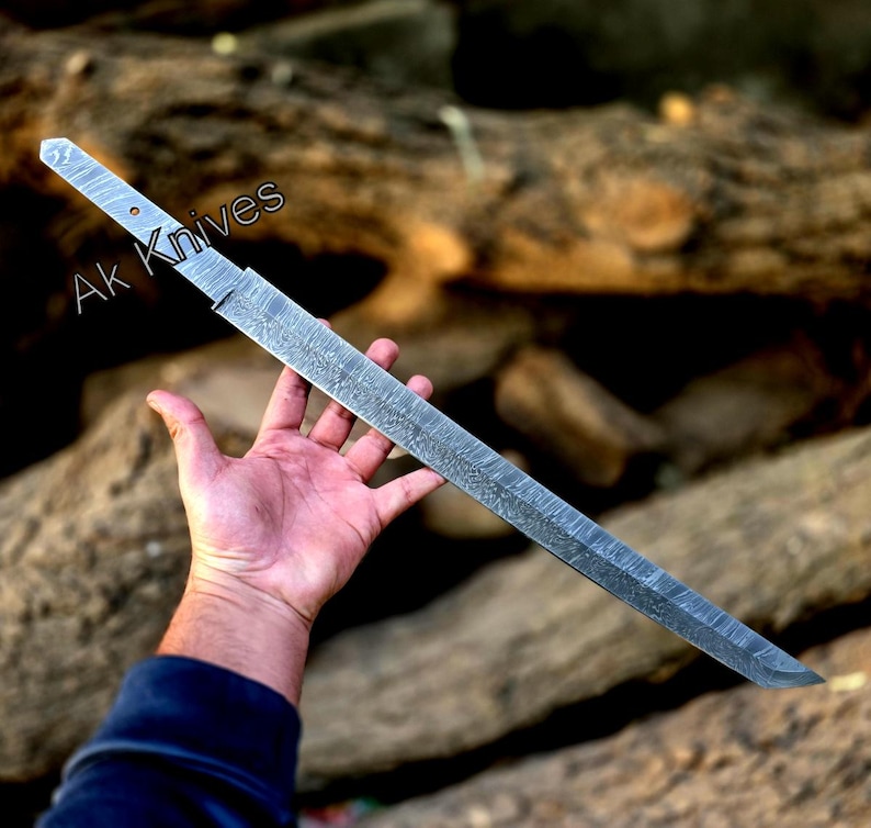Może przedstawiać: Długi, srebrny n&oacute;ż z wzorzystym ostrzem i rękojeścią. Tekst "Ak Knives" jest widoczny na rękojeści. N&oacute;ż trzymany jest w dłoni na rozmytym, naturalnym tle.