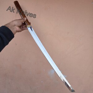 May include: A long, silver blade with a wooden handle and a brass accent. The handle is dark brown. The text "Ak Knives" is visible in the upper left corner. The blade is held at an angle.