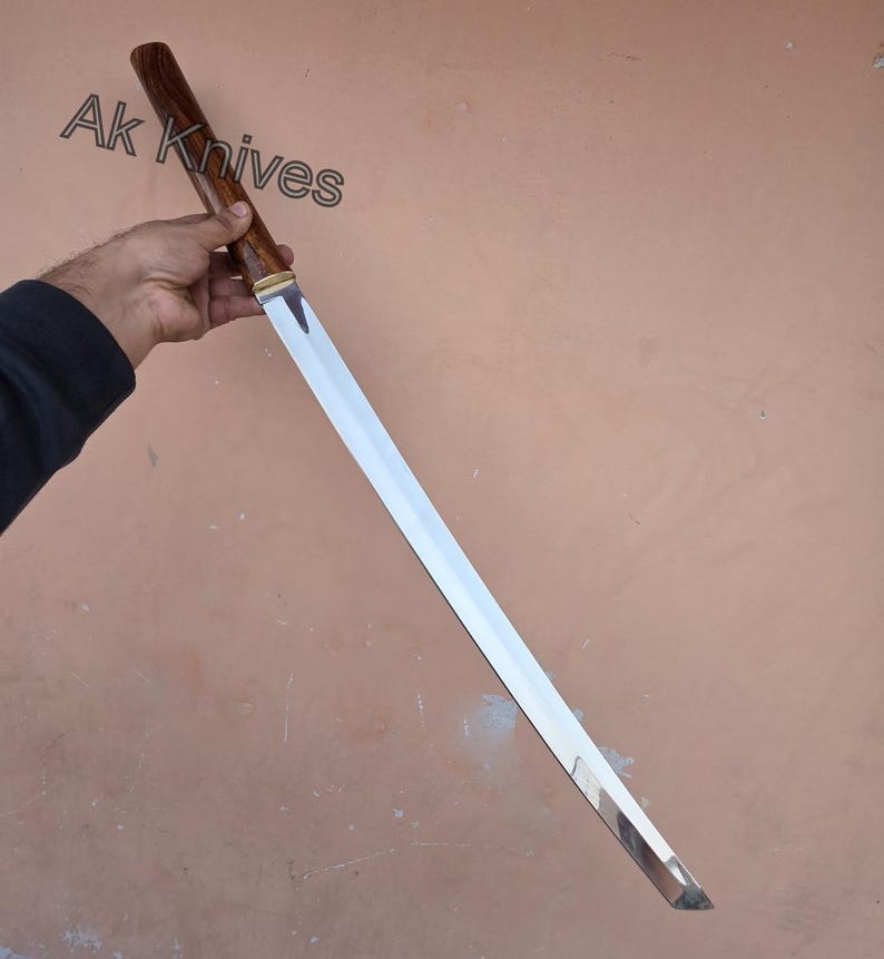 May include: A long, silver blade with a wooden handle and a brass accent. The handle is dark brown. The text "Ak Knives" is visible in the upper left corner. The blade is held at an angle.