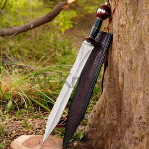 Könnte beinhalten: Ein römisches Gladius-Schwert mit polierter Stahlklinge und Holzgriff mit dunkelbraunem Knauf. Das Schwert ist in einer Lederscheide mit dunkelrotem Innenraum und schwarzem Äußeren verstaut und lehnt an einem Baum.