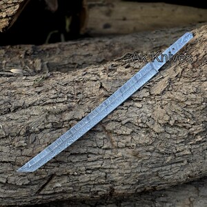 Może przedstawiać: Długie, srebrne ostrze noża z wzorem, spoczywające na teksturowanej drewnianej powierzchni. Na ostrzu widoczny jest napis "Ak Knives". Ostrze ma ostry, kątowy czubek i płaski, prostokątny uchwyt.