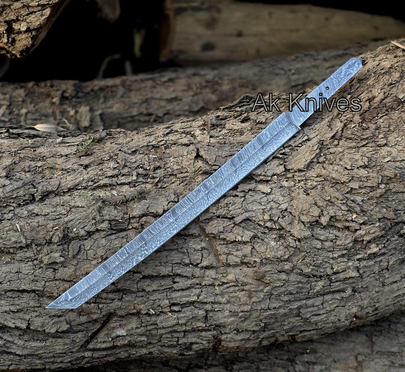 Może przedstawiać: Długie, srebrne ostrze noża z wzorem, spoczywające na teksturowanej drewnianej powierzchni. Na ostrzu widoczny jest napis "Ak Knives". Ostrze ma ostry, kątowy czubek i płaski, prostokątny uchwyt.