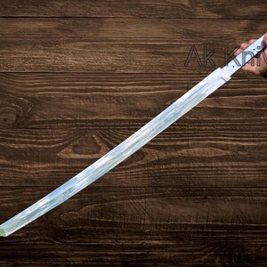 May include: A long, straight sword with a white handle and a reflective blade. The blade has a slight curve and a sharp point. The text "Ak Knives" is visible on the wooden background.