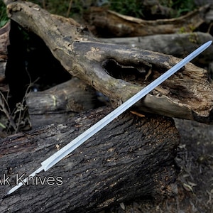 May include: A long, silver sword with a pointed tip rests on a weathered, dark brown log. The sword's blade is straight and appears to be made of metal. The text "Ak Knives" is visible on the log.