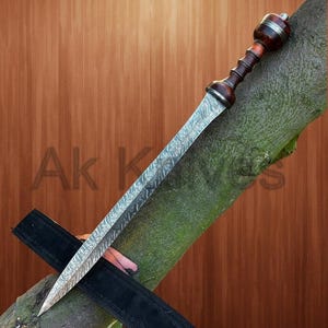 Könnte beinhalten: Ein detailliertes Bild eines Gladius-Schwertes mit gemusterter Klinge und Holzgriff. Der Griff ist dunkelbraun mit silbernen Akzenten. Das Schwert liegt auf einem Ast mit einer schwarzen Scheide. Der Text "Ak Knives" ist sichtbar.