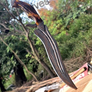 May include: A curved, Damascus steel knife with a brown and tan antler handle. The blade has a distinctive pattern and a sharp point. The knife is designed with a curved blade and a guard. The text "AK Knives" is visible in the background.