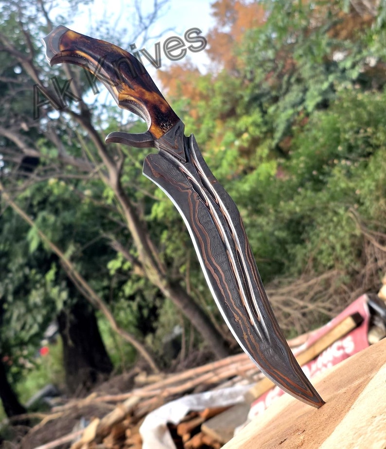 May include: A curved, Damascus steel knife with a brown and tan antler handle. The blade has a distinctive pattern and a sharp point. The knife is designed with a curved blade and a guard. The text "AK Knives" is visible in the background.