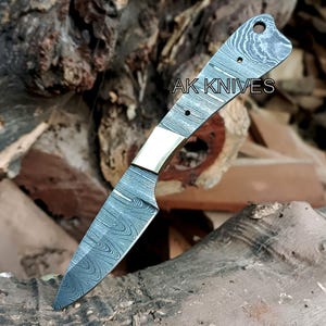 May include: A Damascus steel knife with a patterned blade and handle. The handle has a brass bolster and is marked "AK KNIVES". The knife is set against a backdrop of wood.