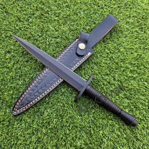 May include: A black dagger with a double-edged blade, resting on a black leather sheath with decorative stitching. The handle is black and ribbed. The dagger is displayed on a green surface.
