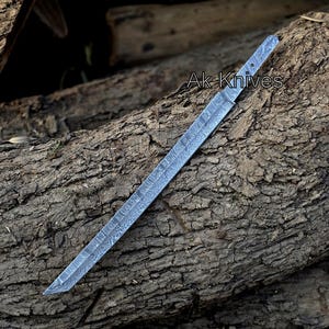 Może przedstawiać: Długie, wzorzyste ostrze ze stali z ostrym ostrzem, spoczywające na teksturowanej drewnianej powierzchni. Ostrze ma unikalny wz&oacute;r, a tekst "Ak Knives" jest widoczny.